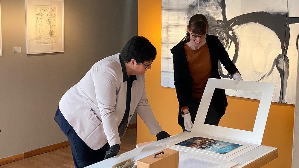 Sabine Verheyen leans forward to look at a print that lays on a table. Another person on her side has lifted a paper frame off the print.   