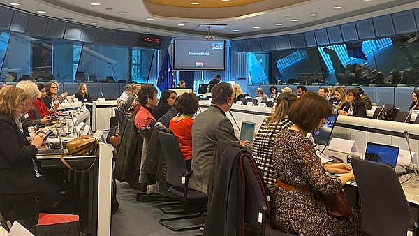 This photograph shows people sitting inside a conference room. Most of them have their backs to the camera and are working on laptops