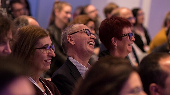 This photograph shows an audience in a dimly lit room. The focus is on three people who are laughung.