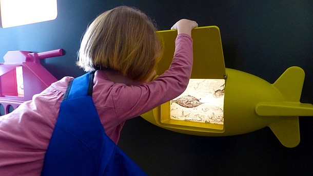  A child is photographed from the back while lifting a flap on a small yellow submarine. Behind the flap there is an illuminated picture.