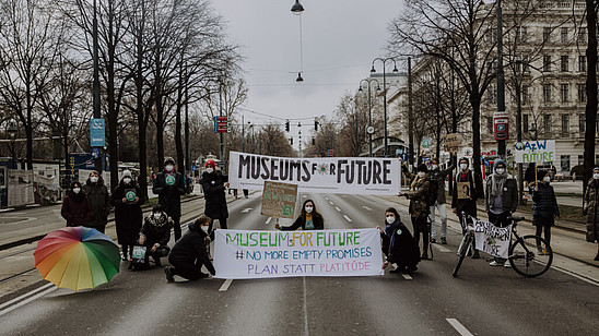 Multiple people are standing and squatting in the middle of a street. They are holding posters and banners. One of them reads "Museums for future".