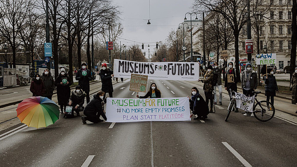  Multiple people are standing and squatting in the middle of a street. They are holding posters and banners. One of them reads "Museums for future".