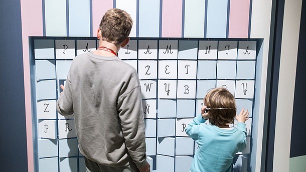  Two children are shown from the back enganging with an exhibition display. They are turning moveable tiles on which letters are printed.