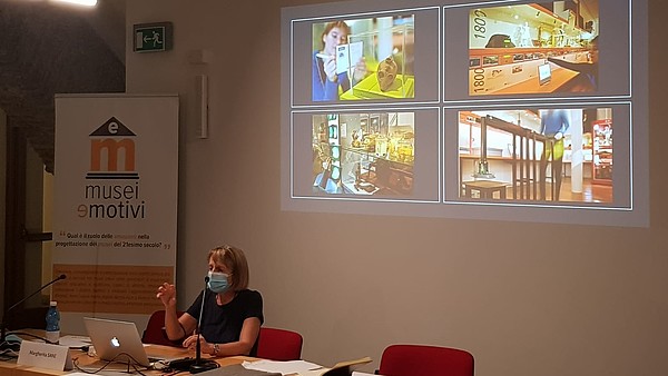 A person is sitting at a desk speaking into a microphone and wearing a mask. On the wall behind them presentation slides are projected with a beamer.