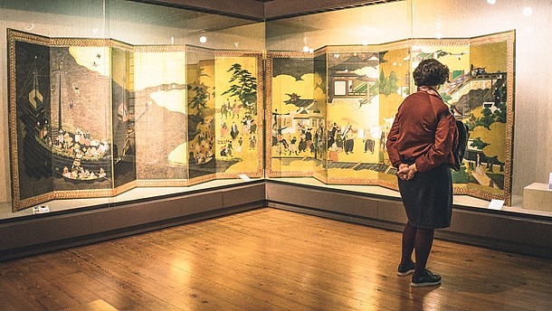  A person with their hands on their back looks at a kind of folded art piece that covers the corner of a room. The art piece is mounted in a glass monter.