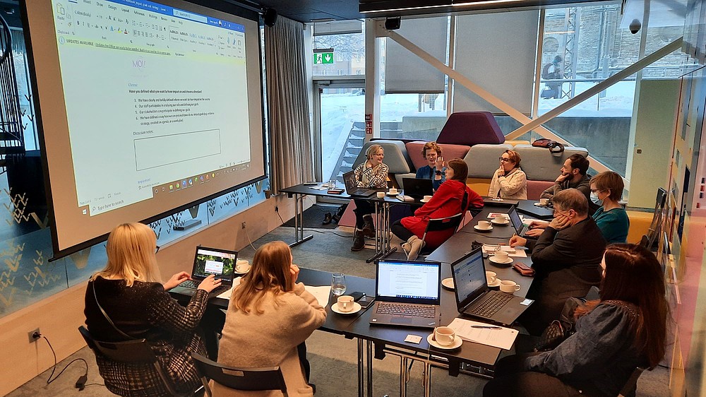 A group of people is sitting in a U-shape with laptops in front of them. A text document is projected on a screen at the front.  