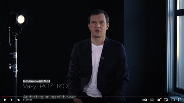  This image is a video still. Vasyl Rozhko sits in a room with dark walls. In the background a spotlight is directed at him.