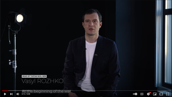 This image is a video still. Vasyl Rozhko sits in a room with dark walls. In the background a spotlight is directed at him.