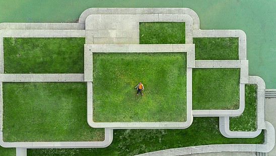 Aerial view of a modern architectural structure made out of concrete rectangles. Inside the rectangles grass is growing and a person is laying down in one of them. 