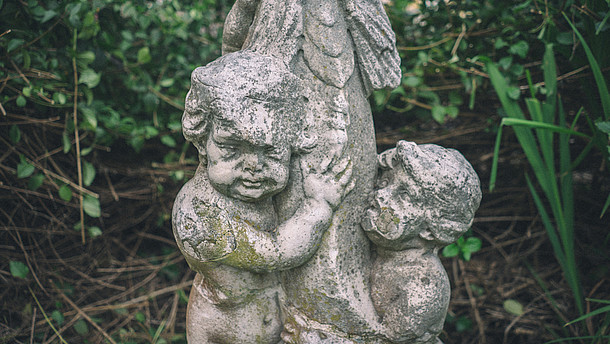  Detail of a stone statue depicting two children. The statue is placed outside and weathered.