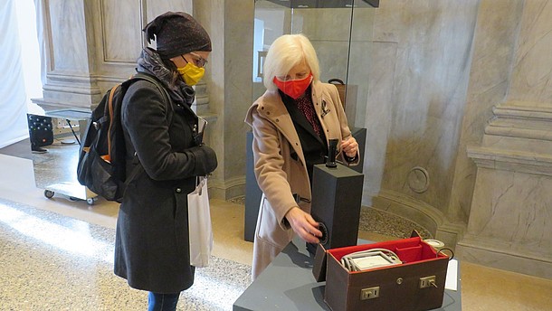  Two people wearing face masks are looking at an object that is placed on a display plinth. 