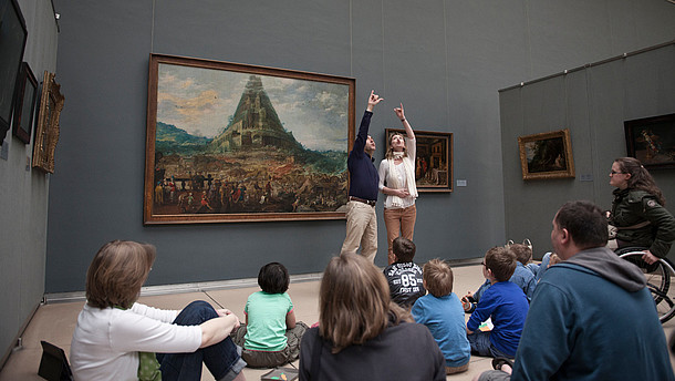  Two guides make a big gesture together to explain a painting to a group of people.