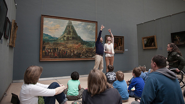 Two guides make a big gesture together to explain a painting to a group of people.