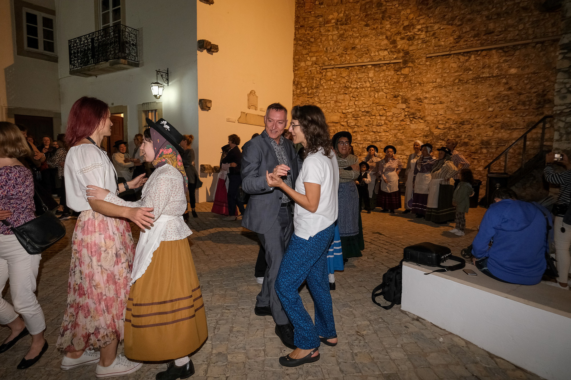 People are dancing in pairs outside in the evening. In the background is an ancient looking wall.  © Image: Jorge Gomes