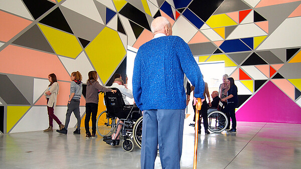 An old man with a cane is standing with his back to the camera in a room with colourful and abstract wall art. In front of him a bunch of people are looking at the art.