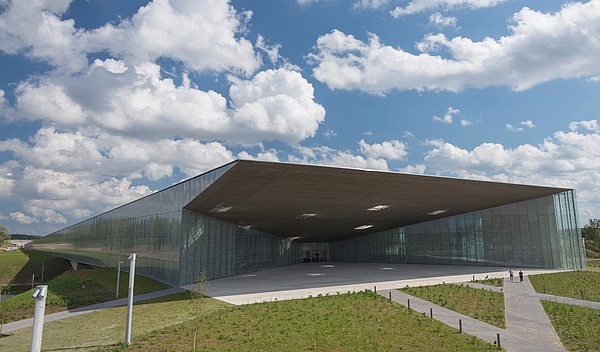 A flat and modern building structure, made mostly out of glass, is surrounded by green nature. Two people are walking towards it.