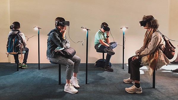 Four people are sitting in a gallery space. All four are wearing goggles and headphones and they are experiencing some kind of virtual reality art.