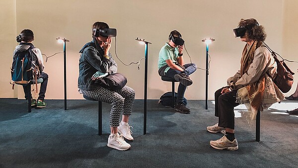 Four people are sitting in a gallery space. All four are wearing goggles and headphones and they are experiencing some kind of virtual reality art.