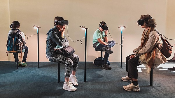Four people are sitting in a gallery space. All four are wearing goggles and headphones and they are experiencing some kind of virtual reality art.