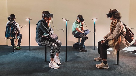 Four people are sitting in a gallery space. All four are wearing goggles and headphones and they are experiencing some kind of virtual reality art.