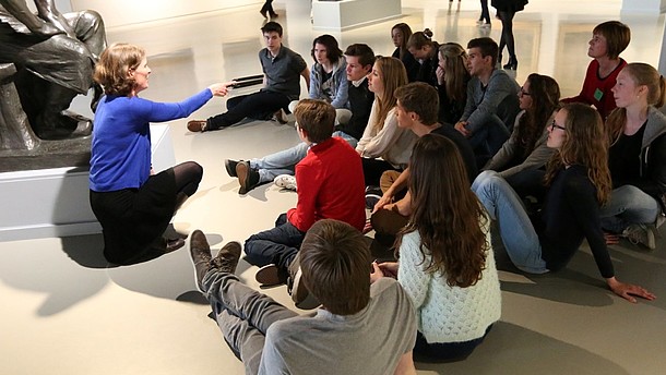 Group of young people are sitting down by a statue in a gallery and listen to a person.  © Image: J. Pierrard