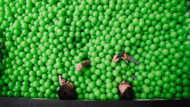  Two people. photographed from bird perspective, sit in a pit of green plastic balls. They are both using their smartphones.
