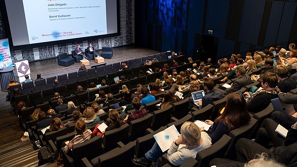 Auditorium full of people looking at two people on the stage. A sign shows that they are discussing EU culture policy. 