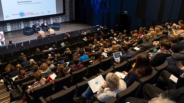 Auditorium full of people looking at two people on the stage. A sign shows that they are discussing EU culture policy. 
