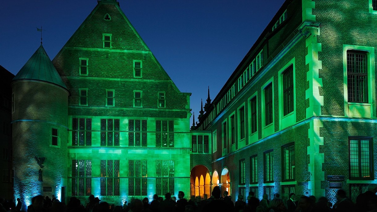© Werner Otto / Alamy Stock Foto An old brick building is lit by green spotlights. In front of it numerous people have gathered to watch it.