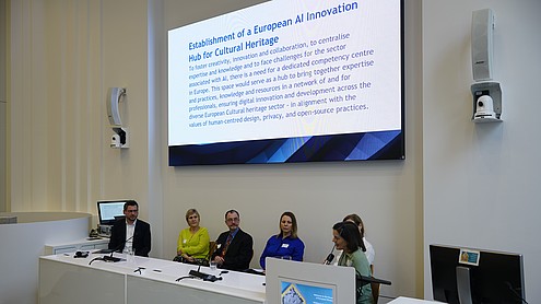 6 persons are sitting behind a desk. One person is speaking into a hand-held microphone. Behind them is an image projected that reads: "Establishment of a European AI innovation hub for cultural heritage - To foster creativity, innovation and collaboration, to centralise expertise and knowledge and to face challenges for the sector associated with AI, there is a need for a dedicated competency centre in Europe. This space would serve as a hub to bring together expertise and practices, knowledge and resources in a network of and for professionals, ensuring digital innovation and development across the diverse European Cultural heritage sector - in alignment with the values of human-centred design, privacy, and open-source practices."   © Image: Peter Vand der Plaetsen