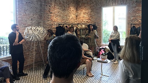People are spread out in a fashion showroom and studio, both sitting and standing, while listening to a presentation by a fashion designer.  