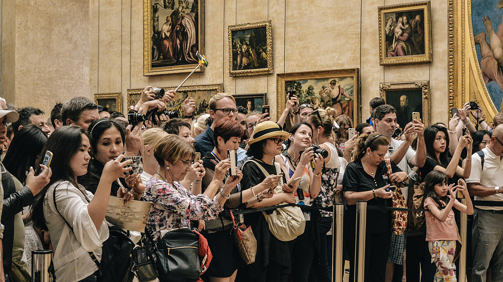 In this photo a big group of tourists stand behind a barrier rope inside a museum and take photos. The artwork which they photograph is not shown..  © Image: Alicia Steels