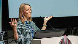 Woman standing behind a podium while giving a presentation is holding her hands up as if she is asking a question.