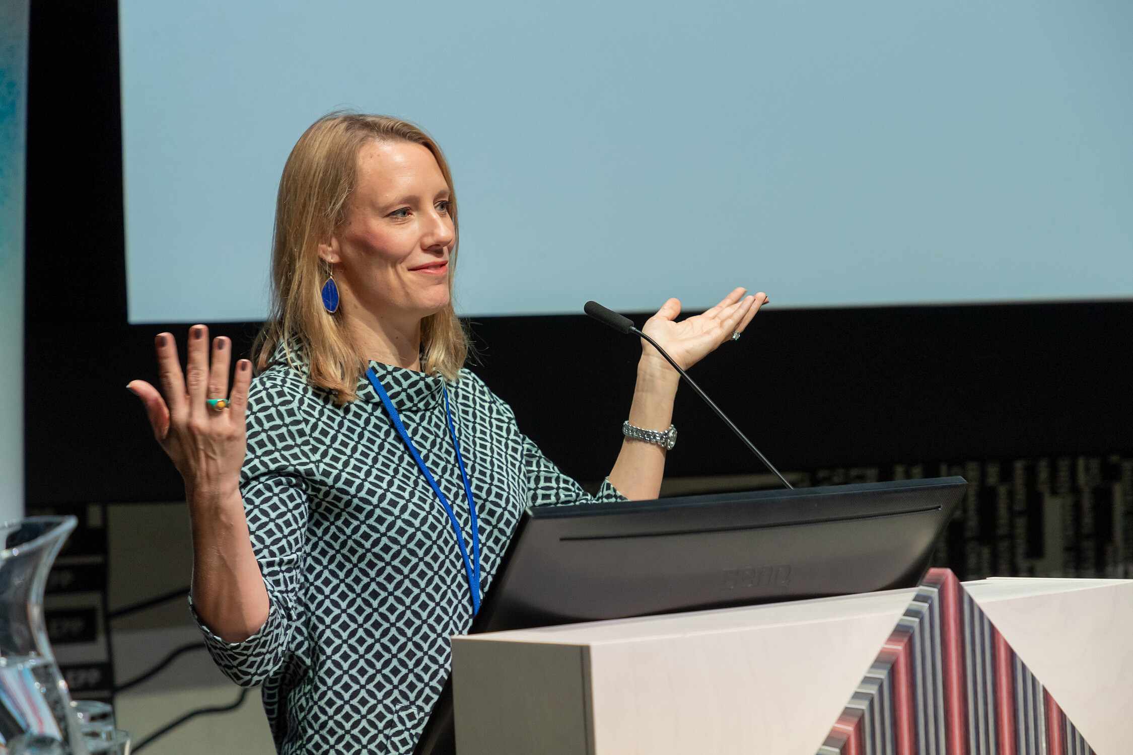 © Image: Arp Karm Woman standing behind a podium while giving a presentation is holding her hands up as if she is asking a question. © Image: Arp Karm