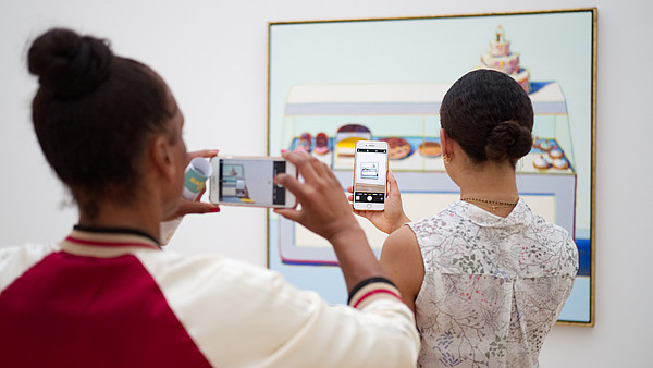 Two people have their backs to the camera as they both take a photo of a painting hanging on the wall. The person in the back is taking her photo of the painting through the phone screen of the person of the person standing in front of her.