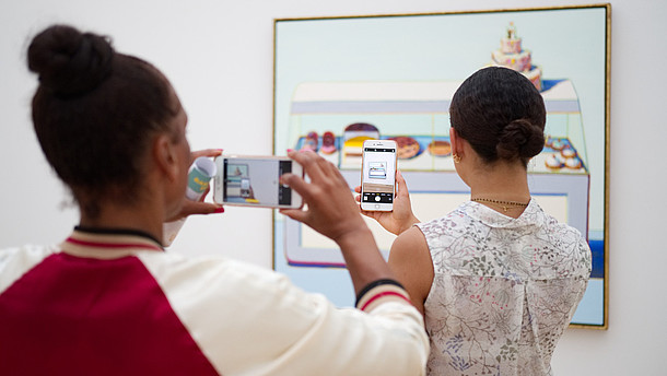  Two people have their backs to the camera as they both take a photo of a painting hanging on the wall. The person in the back is taking her photo of the painting through the phone screen of the person of the person standing in front of her.