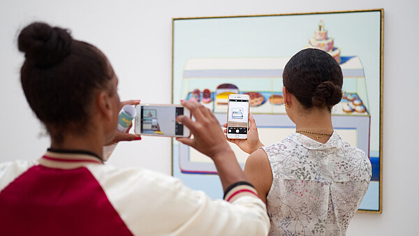 Two people have their backs to the camera as they both take a photo of a painting hanging on the wall. The person in the back is taking her photo of the painting through the phone screen of the person of the person standing in front of her.