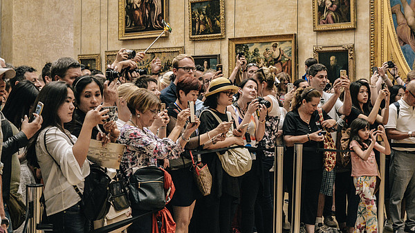 A large group of tourists stand behind a rope fence inside a museum to take photos of something outside of the picture.