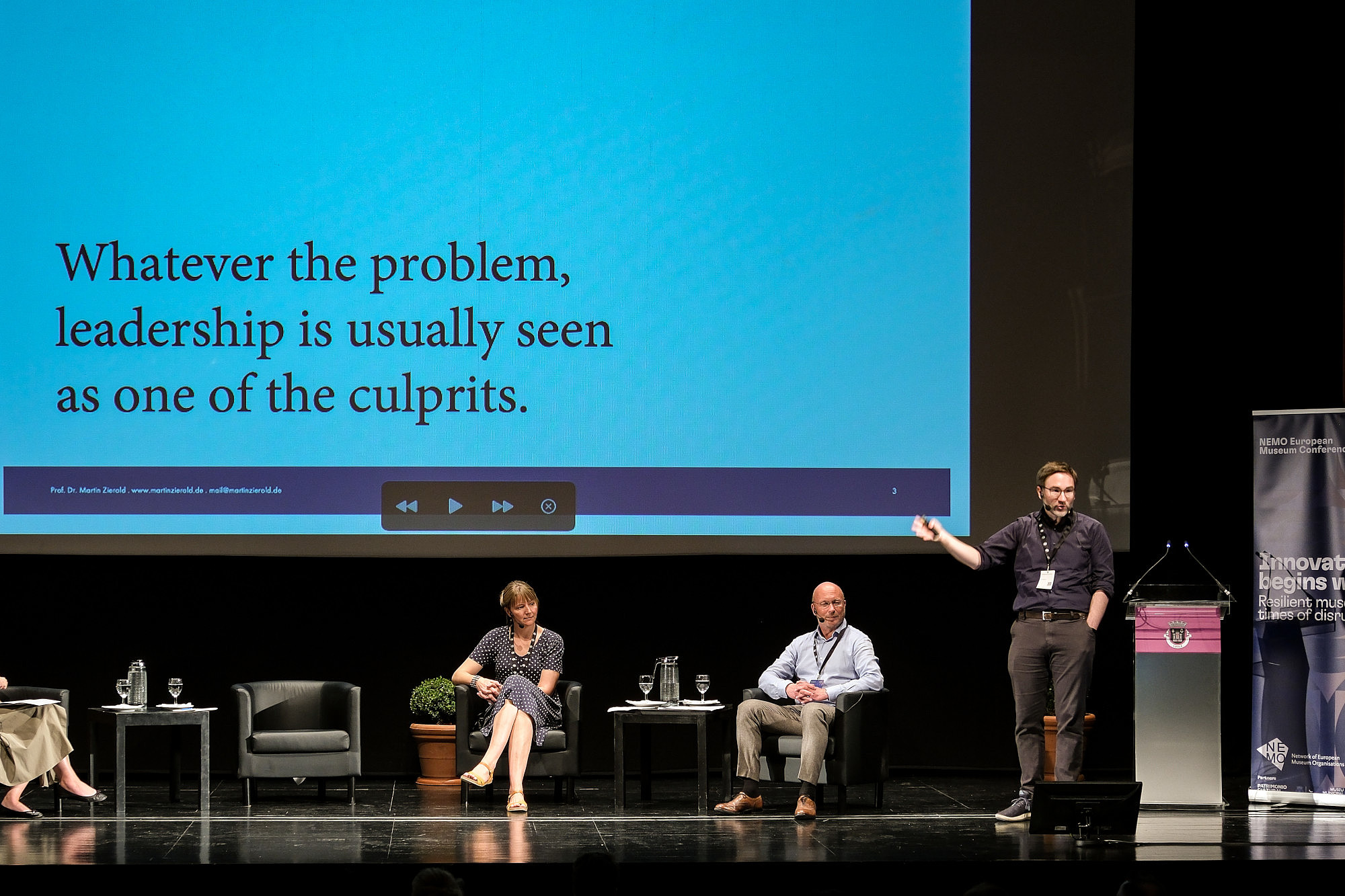 Three people are sitting on a stage. Another person is standing, gesturing with their arms and talking. In the background, presentation slides are projected to a screen. The headline on the slide reads: "Whatever the problem, leadership is usually seen as one of the culprits."  © Image: Jorge Gomes