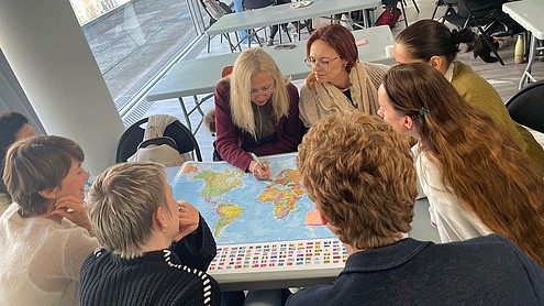 Six people are seated around a table leaning over a map of the world. With pens they are mapping out recent travel in the last year.   