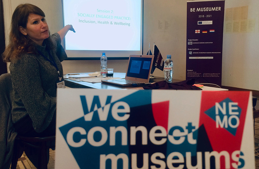 Person is sitting down whilst giving a presentation. She points at the presentation that is projected on a screen. A foreground someone is holding up a postcard with NEMO's logo that reads "We connect museums"   © Image: Lana Karaia