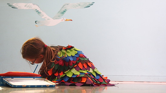 A child dressed in a colourful, eccentric robe is sitting on the floor writing.