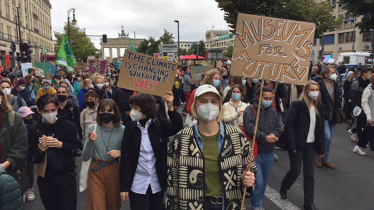  This picture shows a protest with many people marching together. All of them are wearing face masks. In the foreground a person holds up a poster stating "Museums for Future".