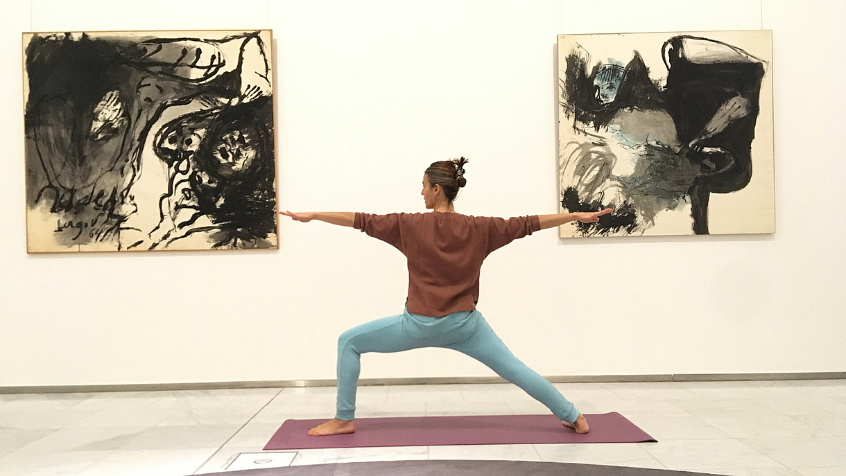  A person is photographed from the back while doing a yoga pose inside a museum space. 