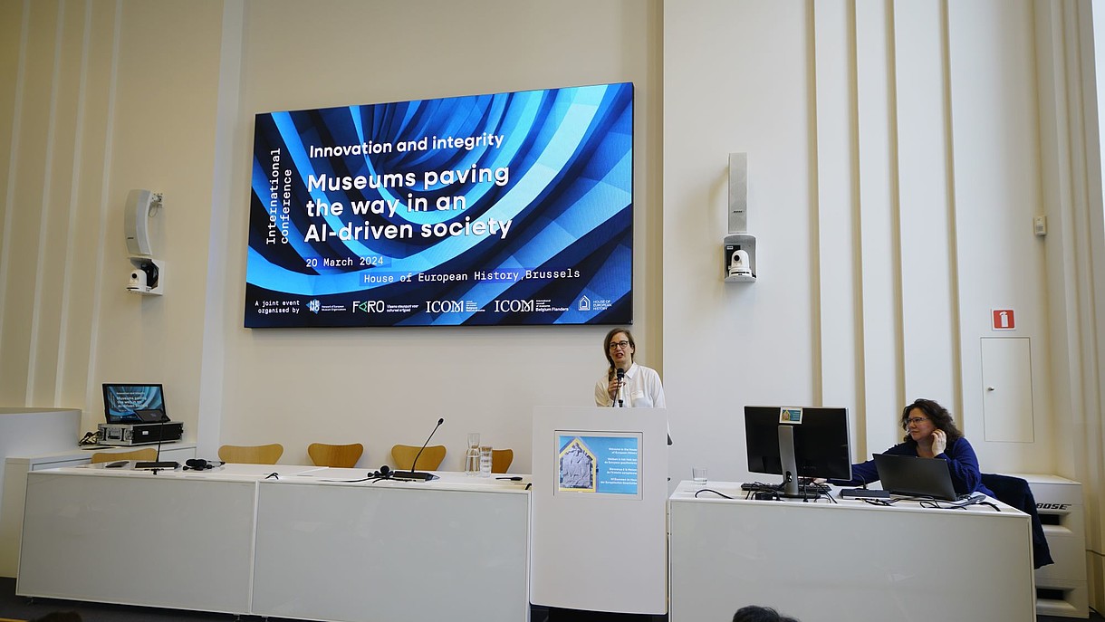© Image: Peter Van der Plaetsen A person is standing behind a podium talking into a handheld microphone. Behind her there is a projected image with the name of the conference "Innovation and Integrity: Museums paving the way in an AI-driven society".