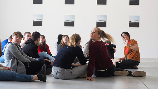 A group of young people are sitting on the floor in an exhibition space. A museum educator is speaking to them.