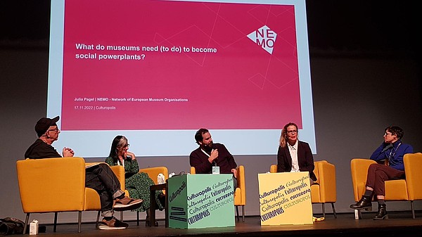 This photograph shows five people sitting on a stage. They are wearing headphones and one of them is talking while looking at the audience. In the background presentation slides are projected.