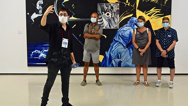  A man is taking a selfie of himself and three other people standing behind him in front of a big painting. They are all wearing face masks. 