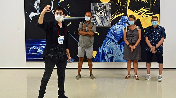 A man is taking a selfie of himself and three other people standing behind him in front of a big painting. They are all wearing face masks. 
