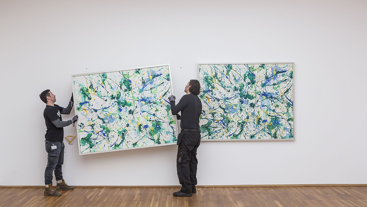 © Image: David von Becker © Image: David von Becker Two people are hanging a painting on a wall next to a painting that already is hung up.